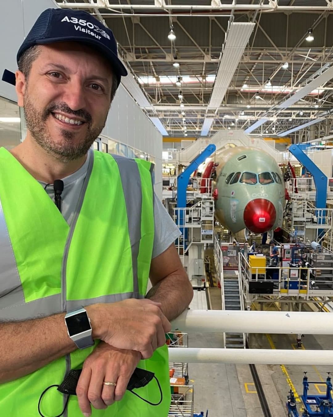 Lito Sousa in a safety vest and an A350 visitor cap, smiling inside an aircraft assembly hall, with an airliner fuselage behind him.